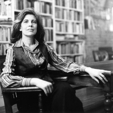 Susan Sontag sitting in wooden chair at wooden table with bookshelves in the background