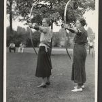 Two women in a park hold bows and arrows, ready to shoot