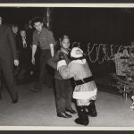 boy looking surprised when approached by chimpanzee dressed in santa suit and adults look on