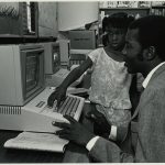 A child teaches a man to use the computer at the library.