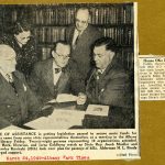 A group of men and women gather in the library in this sepia-toned newsprint photograph.