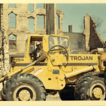 A man in a yellow bulldozer looks back incredulously at his colleague. Behind them is a destroyed building.