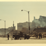 A national guard truck and guard members stand on the street in front of burnt-out buildings.