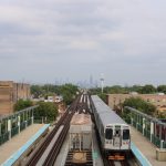 A daytime photo of the CTA green line's Laramie stop. One train is at the station. The fog-enshrouded buildings of downtown Chicago loom in the horizon.