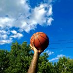 A tattooed arm lifts a basketball into the air. The sky is bright blue with white clouds, and sun glints off the ball.