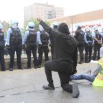 Viewed from behind, a Black man in all black kneels and raises his fist. In front of him, a line of police in riot gear stand with their backs to him,