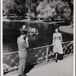 A man taking a photograph of a woman on the walkway above the water in the Boston Public Garden. ca. 1935–1955]