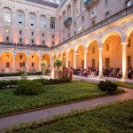 Guests are gathered behind the pillars in seats in the courtyard, while a group of people, including the bride and groom, stand in the courtyard, facing the guests