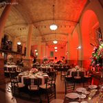 Photo of the Guastavino Room set up with tables and chairs, tablecloths, plates, utensils, and flower arrangements. There is also red lighting.