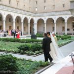 Photo of a bride being walked down the aisle in the McKim courtyard. Opposite her there are guests seated. In front of them is the groom, who is also facing the fountain.