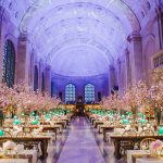 Long view of Bates hall decorated with white and gold tablecloths on the tables, with individual flower arrangements at each seat, as well as large pink flower arrangements at each table