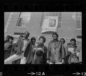 Positive image of a photographic negative showing nine African American standing in front of a building displaying a handmade banner with the partial words Malcol_ Univers_ displayed.