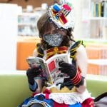 A young person wearing a tiny top hat, skirt, and gloves reads a comic book on a couch at Central Library