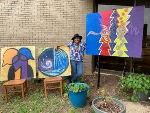 Image is of a female artist wearing a hat posing with 3 of her colorful murals
