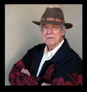 Image is of author Thomas King, smiling at the camera and wearing a hat with a colorful, decorative band
