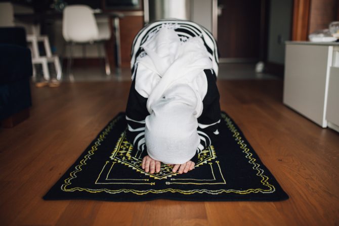 photograph of a woman in her room or apartment bent on a prayer rug