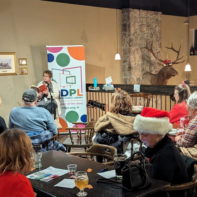Library staff member reading a book with a red cover while sitting on a tall chair in front of a large RDPL banner beside the railing on the top floor at Red Hart Brewing.