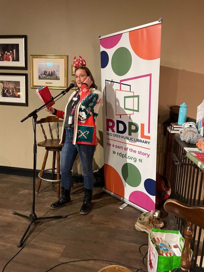 Library staff member wearing a Christmas sweater and reindeer ears, reading a book with a red cover while standing at a microphone in front of a large RDPL banner beside the railing on the top floor at Red Hart Brewing.