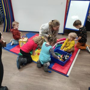 Five small children and one adult playing on a colourful mat with toys
