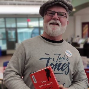 Red Deer's Mayor wearing a newsboy cap and glasses, holding a book with a red cover and wearing a Read for 15 pin