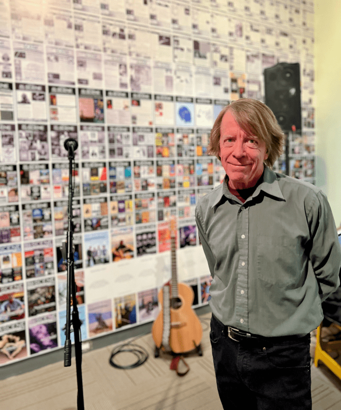 Carl Stretton with a guitar and microphone in the background, and the whole back wall is covered with Red Deer Scene magazine covers.
